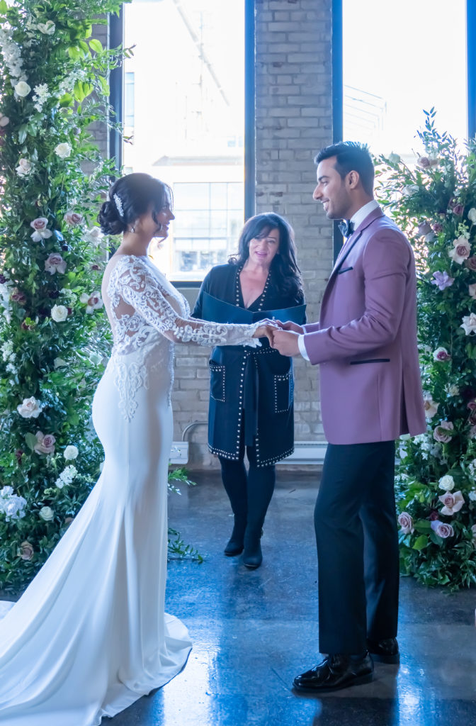Couple holds hands with officiant under a floral arch with white and pink roses  | Weddings & Events by Cheryl Munro | Toronto Wedding Planner